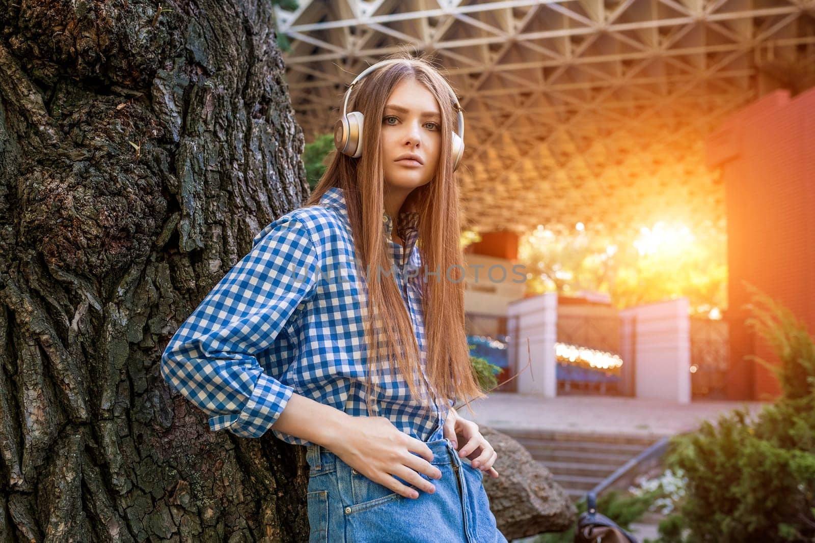 Young woman cute brunette girl standing near tree on headphones, sun light flares of rays, looking at the camera, on summer outdoor copy space background