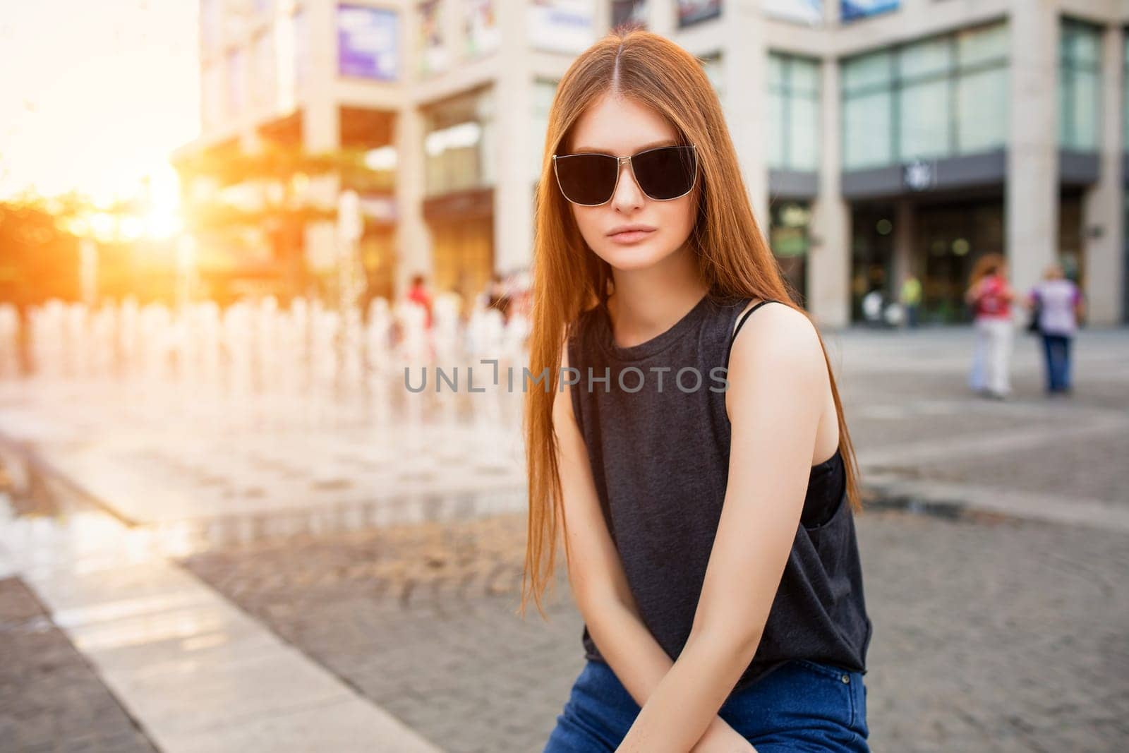 Fashion portrait of beautiful young woman in sunglasses, with view modern city under the warm rays of sun