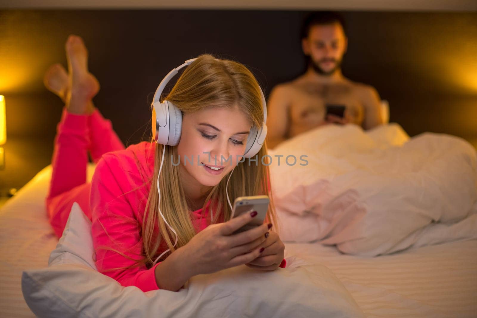 Young happy women on white bed using mobile phone with headphones, boyfriend on background. Concept about technology and people