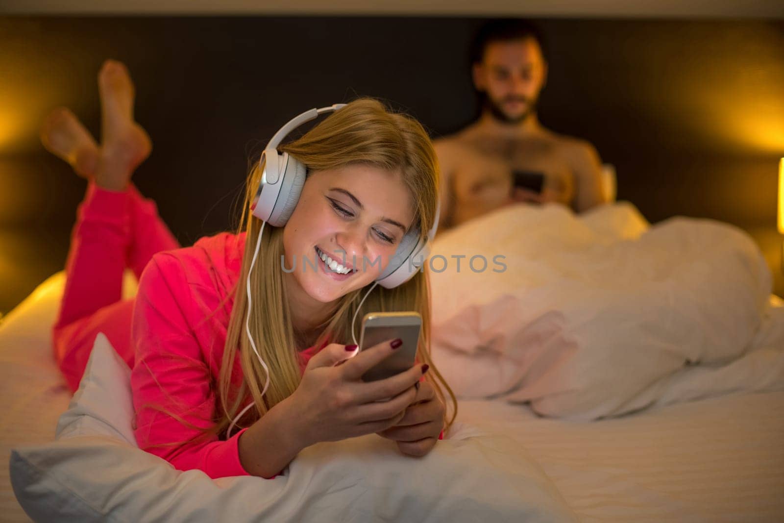 Young happy women on white bed using mobile phone with headphones, boyfriend on background. Concept about technology and people