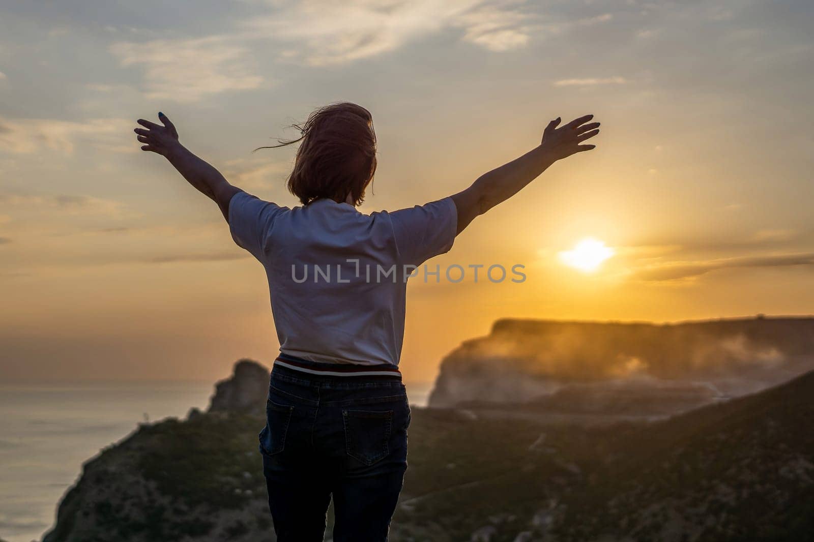 Happy woman on sunset in mountains. Woman standing with her back on the sunset in nature in summer with open hands. Silhouette. by Matiunina