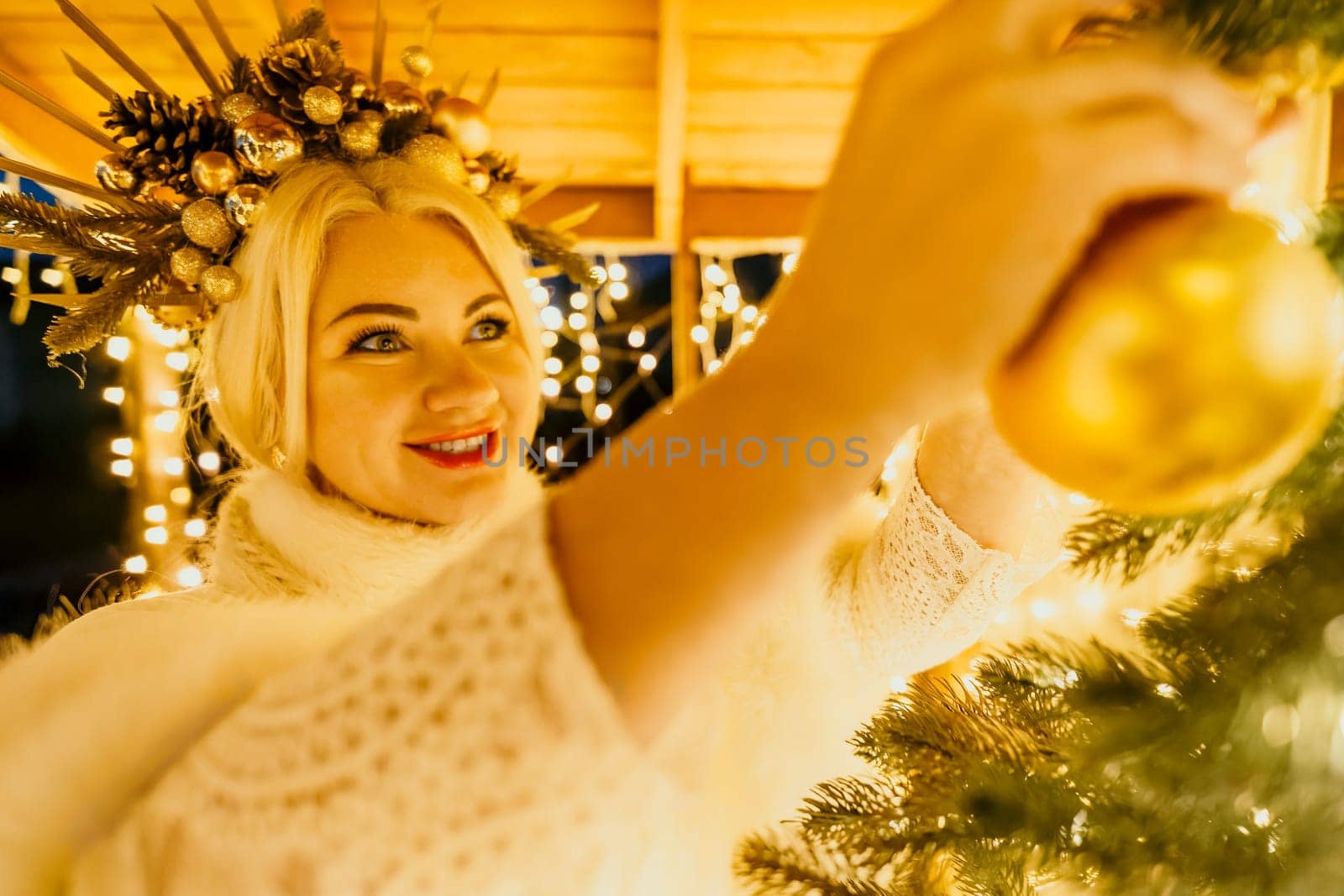A blonde woman in white dress and a crown of gold ornaments decorate Christmas tree with gold ornaments and lights. The tree is decorated with gold balls and is lit up with lights. by panophotograph