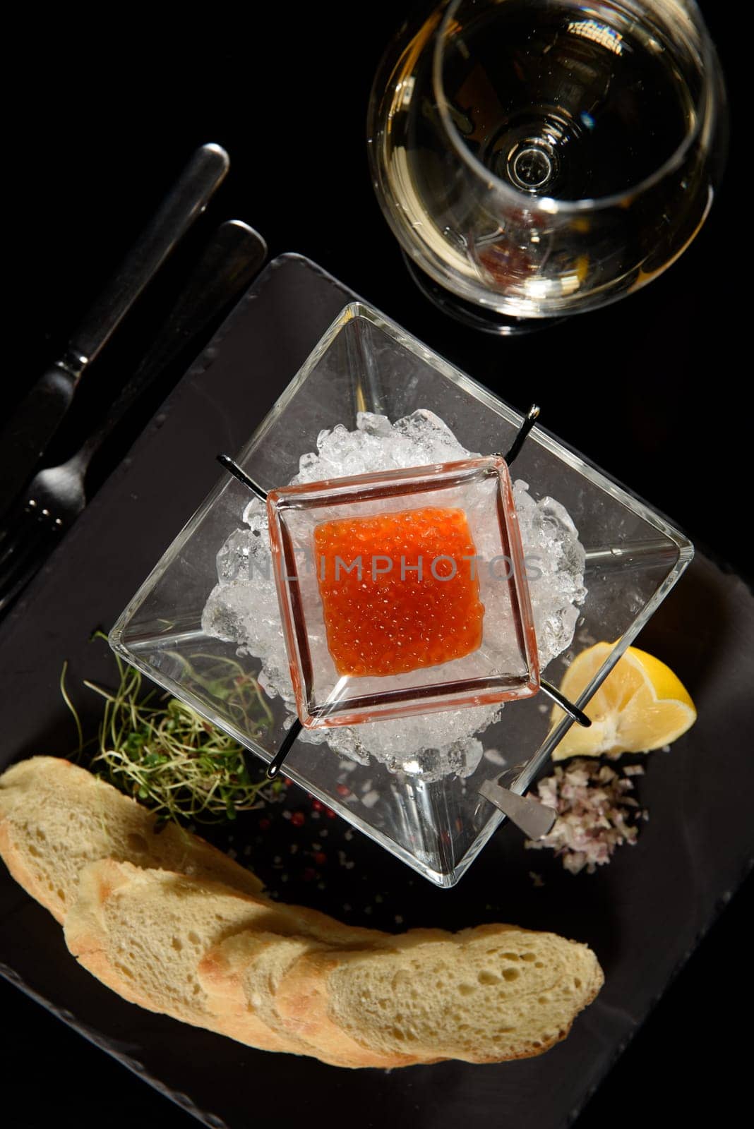 salmon fish caviar, on ice, with croutons and butter, on a transparent dish with a glass of red wine on a dark background