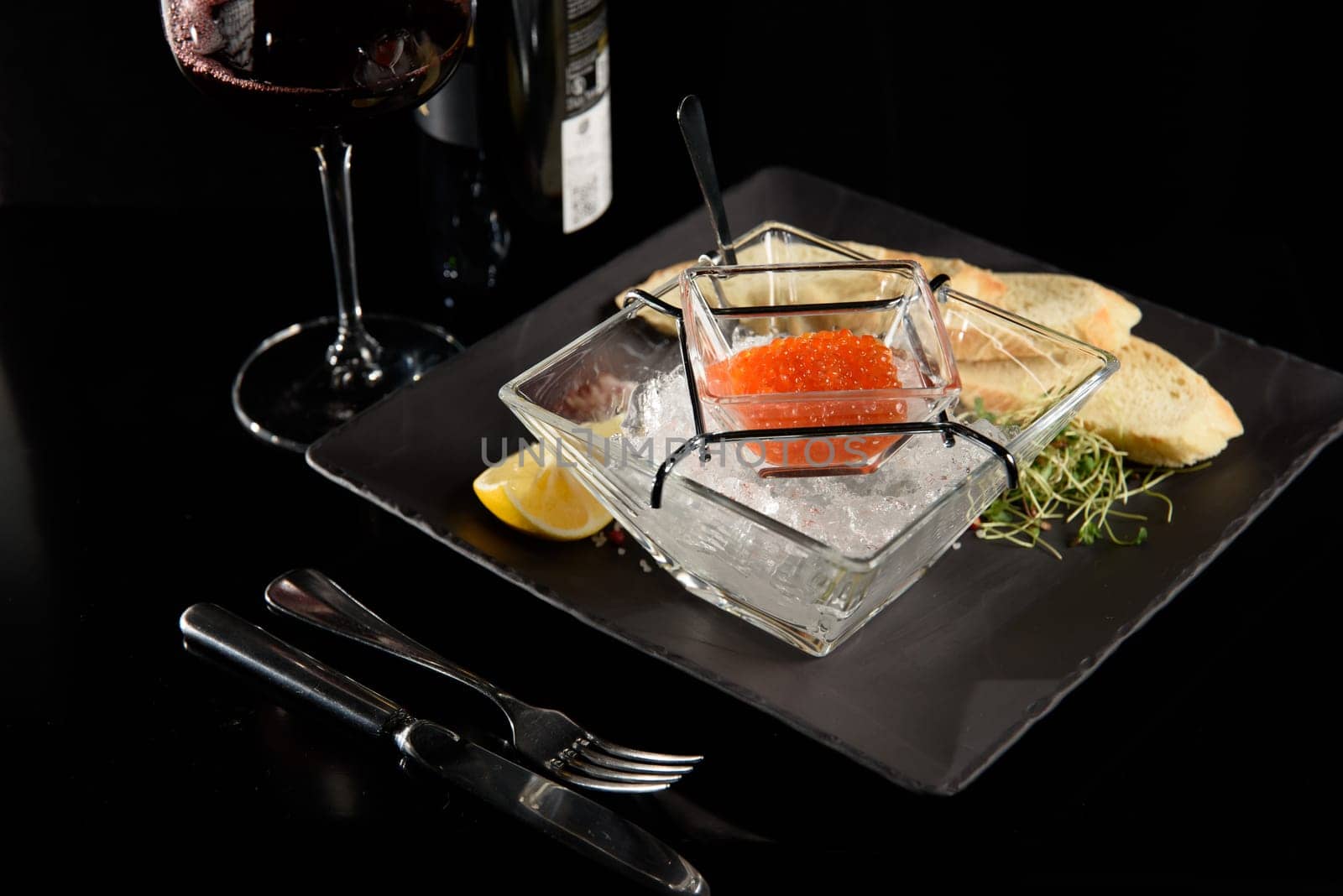 salmon fish caviar, on ice, with croutons and butter, on a transparent dish with a glass of red wine on a dark background