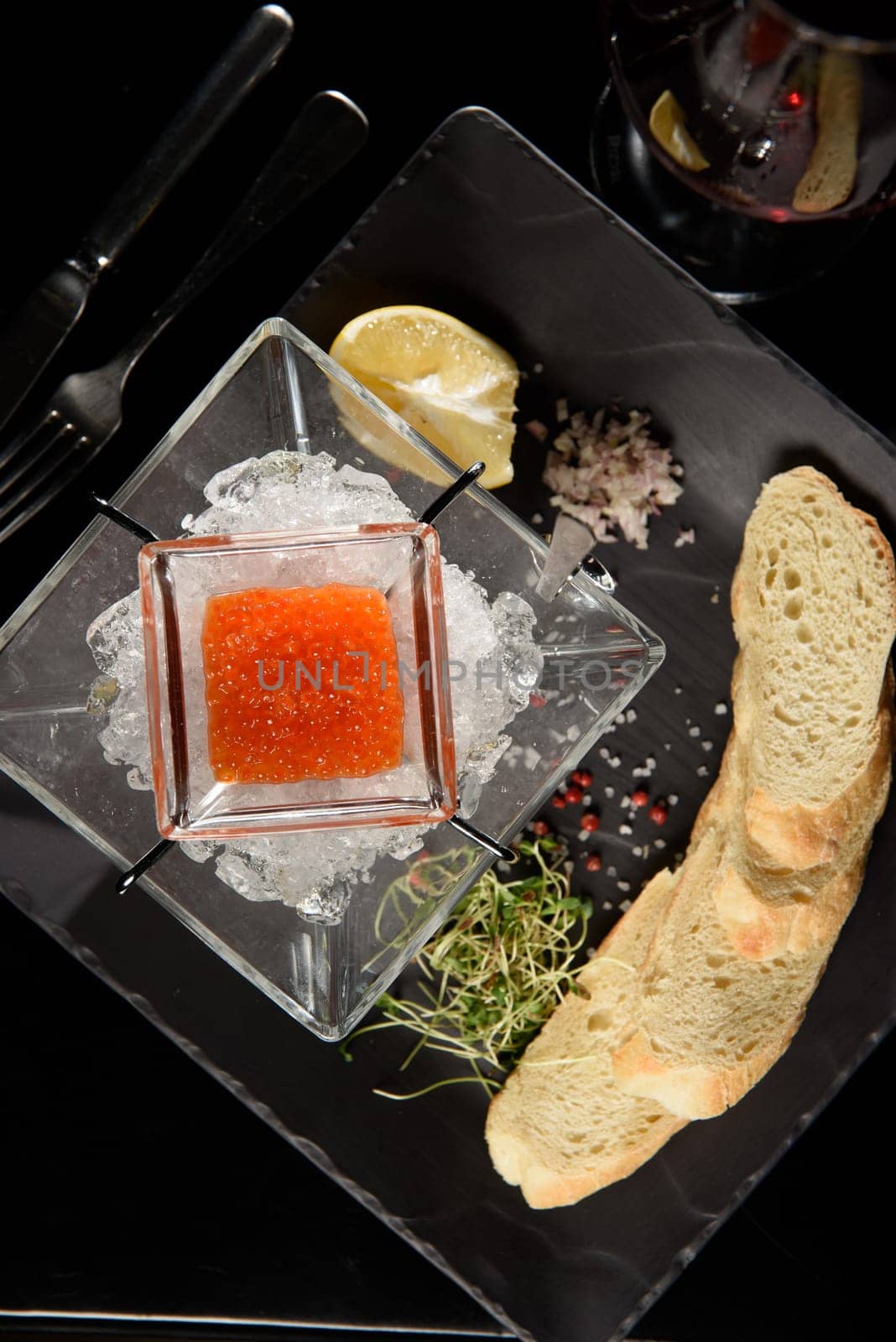 salmon fish caviar, on ice, with croutons and butter, on a transparent dish with a glass of red wine on a dark background