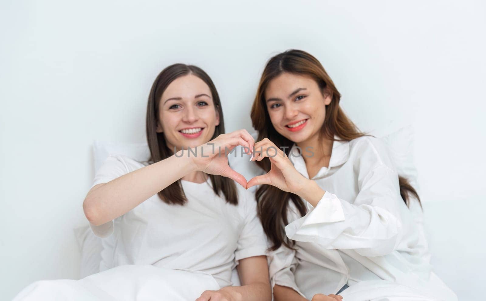 Young Asian lesbian couple sitting on bed during happy resting time, hand made heart symbol. Be happy and relaxed in the bedroom..