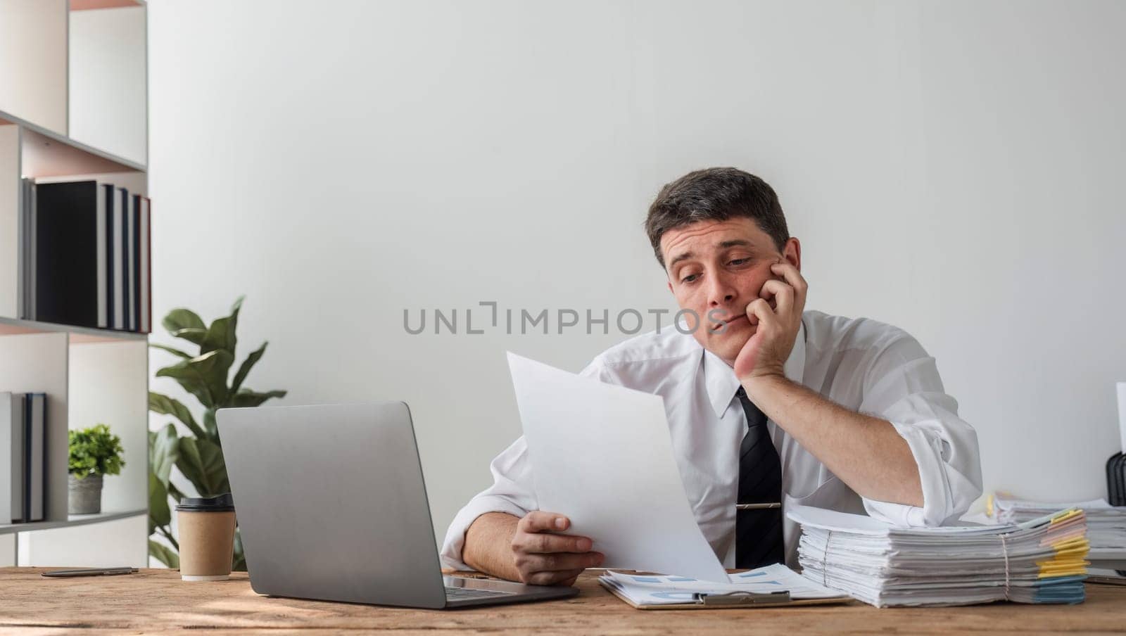 Unhappy young businessman feeling bored and stressed at work looking at laptop with hopeless expression while sitting in office by wichayada