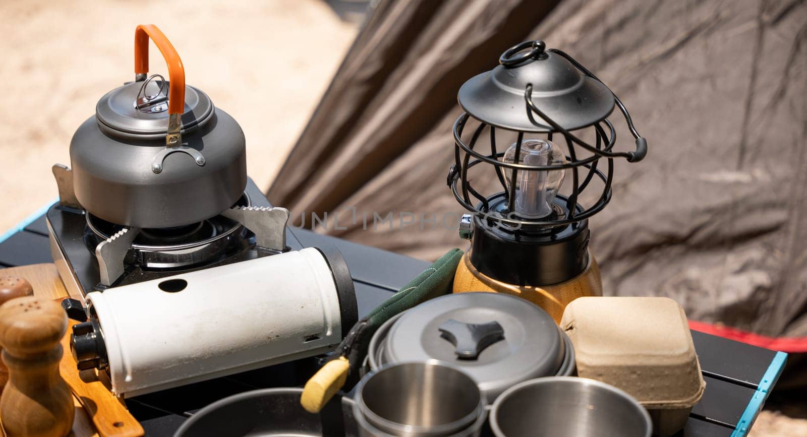 Cooking gear for a tranquil beach campsite, kettle, pot, pan, gas stove, flashlight, and camera set on a table by the tent. Enjoy a serene camping experience amidst nature.