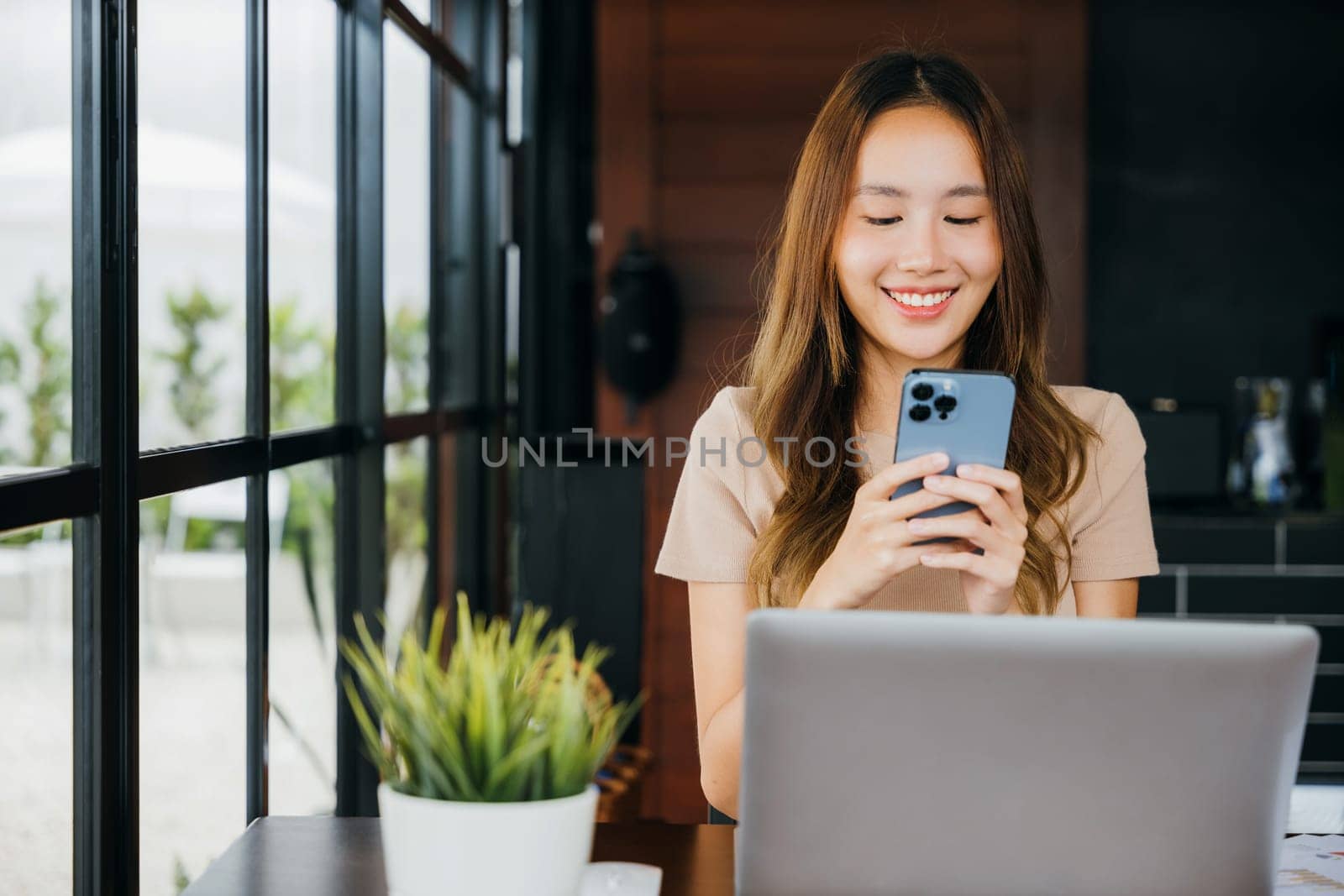 Happy lifestyle female smiling using smartphone in coffee shop, young business woman working with laptop computer she holding smart mobile phone at cefa for texting messages