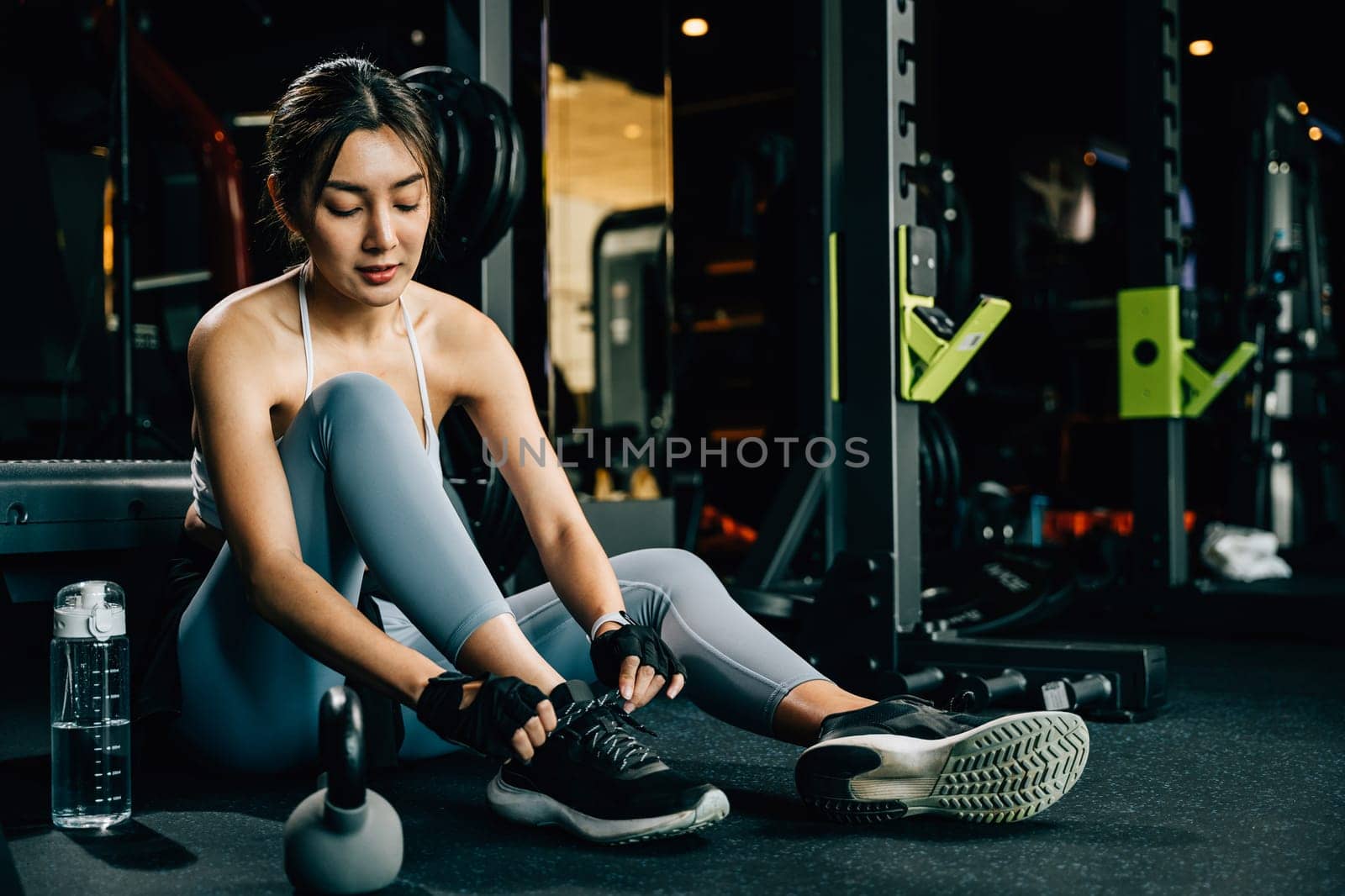 Young woman tying the shoelace of her running shoe on the gym floor. The shot captures her fit and toned legs, showcasing her dedication to fitness.