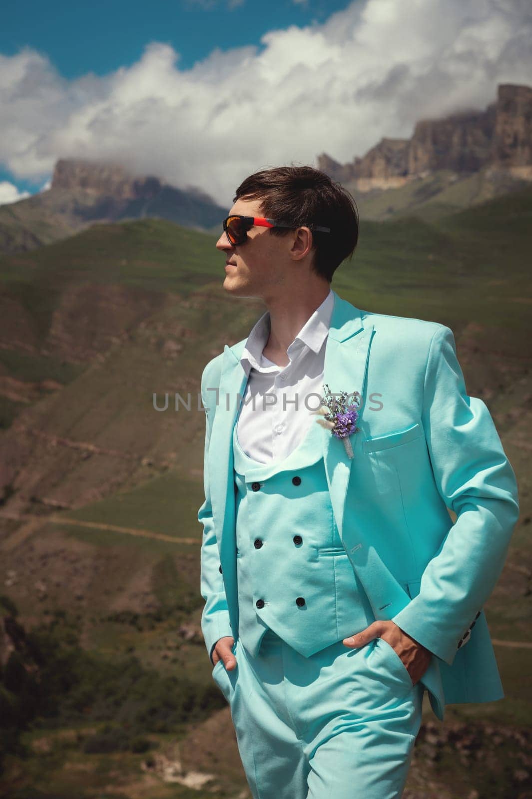 Fashionable portrait of the groom waiting for the bride. stylish groom in sunglasses and a turquoise suit stands alone thoughtfully against the backdrop of a mountain by yanik88