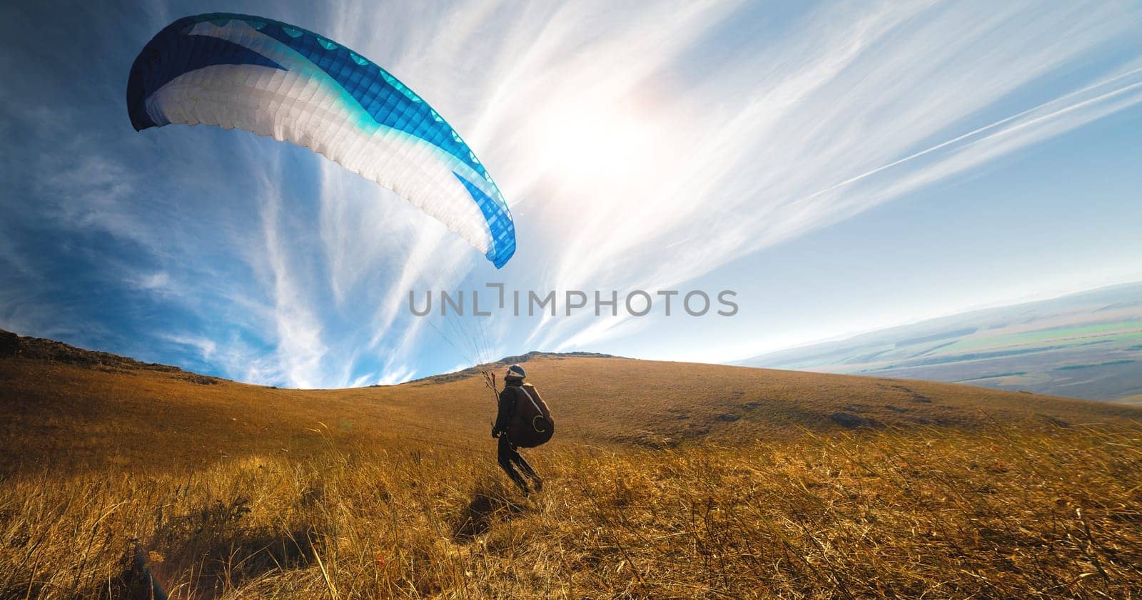 Paragliders take off from a yellow meadow, a man's legs open from the ground, taking off with a parachute upward. Panoramic view of the hills and nature.