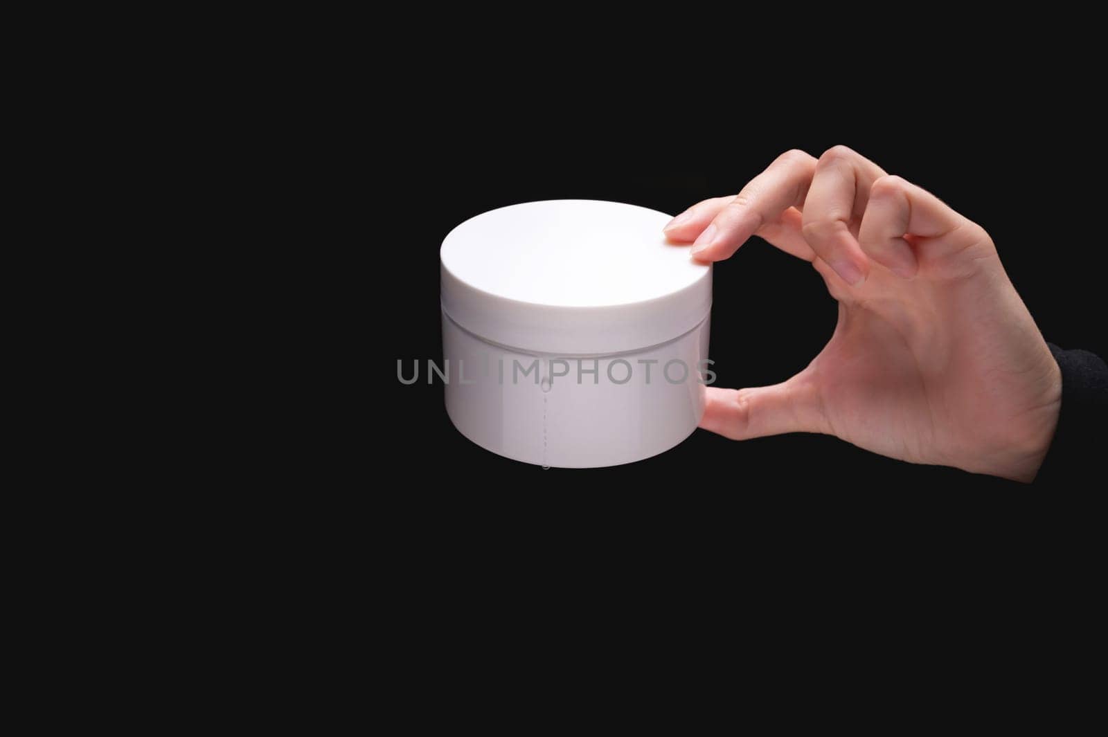 White cosmetic packaging mockup. A woman's hand holds a round jar of body cream on a black background. Beauty industry concept. Close-up by yanik88
