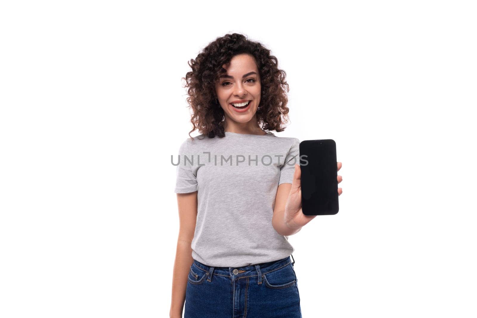well-groomed caucasian young brunette woman with curly hair shows smartphone screen on white background with copy space.