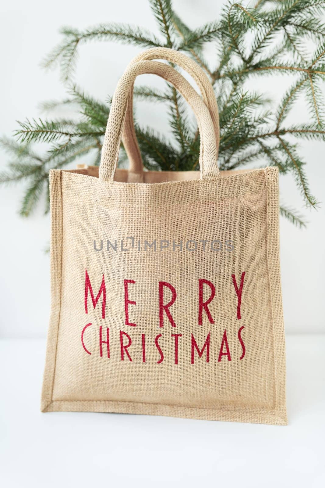 This is a photo of a burlap tote bag with Merry Christmas written in red on it, sitting in front of a small Christmas tree. by sfinks