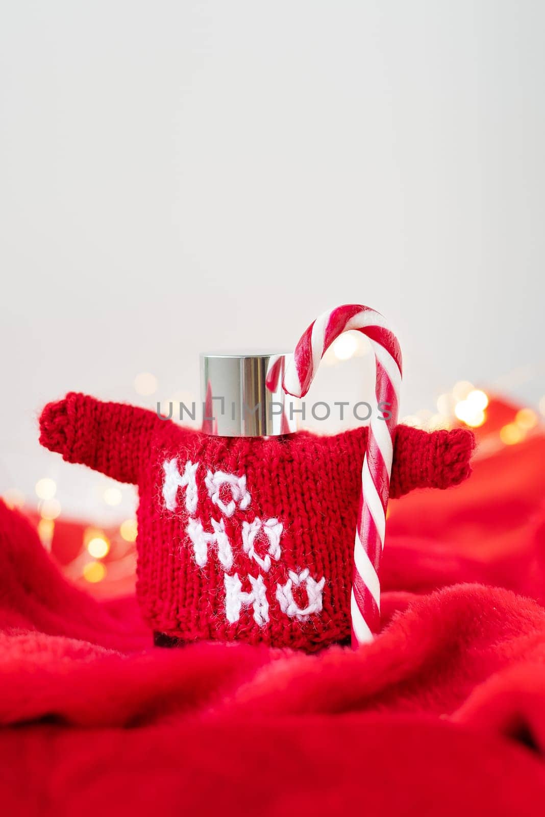 This is a. photo of a red sweater with Ho Ho Ho written on it, wrapped around a silver flask with a candy cane sticking out of it. The background is blurred with Christmas lights and a red blanket