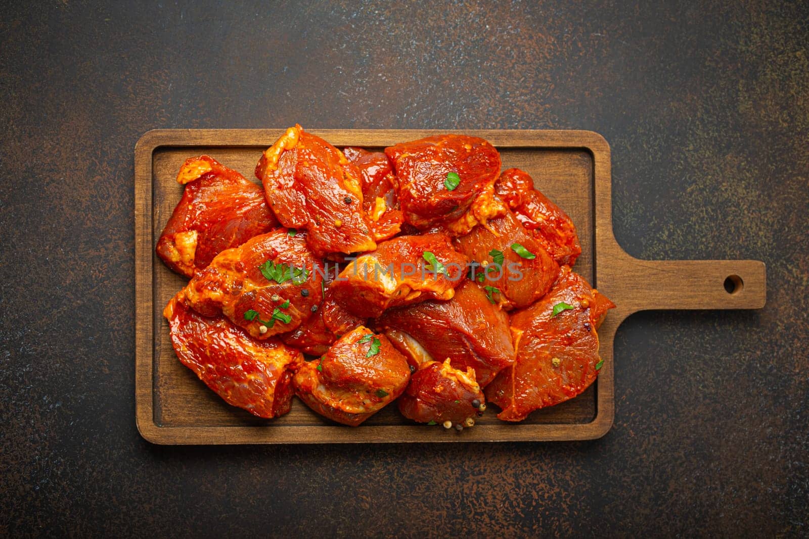 Raw uncooked chopped pieces of pork marinated with seasonings and parsley on wooden cutting board top view on dark rustic background. Cooking meal with marinated pork fillet pieces.