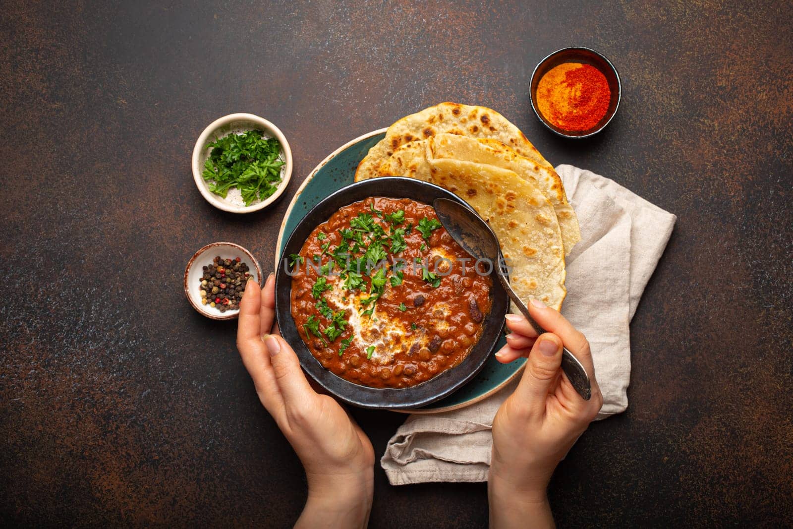 Female hands holding a bowl and eating traditional Indian Punjabi dish Dal makhani with lentils and beans served with naan flat bread, fresh cilantro on brown concrete rustic table top view by its_al_dente