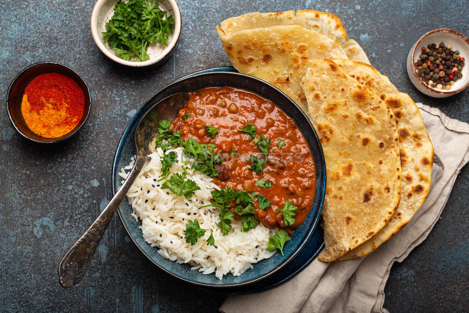 Traditional Indian Punjabi dish Dal makhani with lentils and beans in black bowl served with basmati rice, naan flat bread, fresh cilantro and spoon on blue concrete rustic table top view.