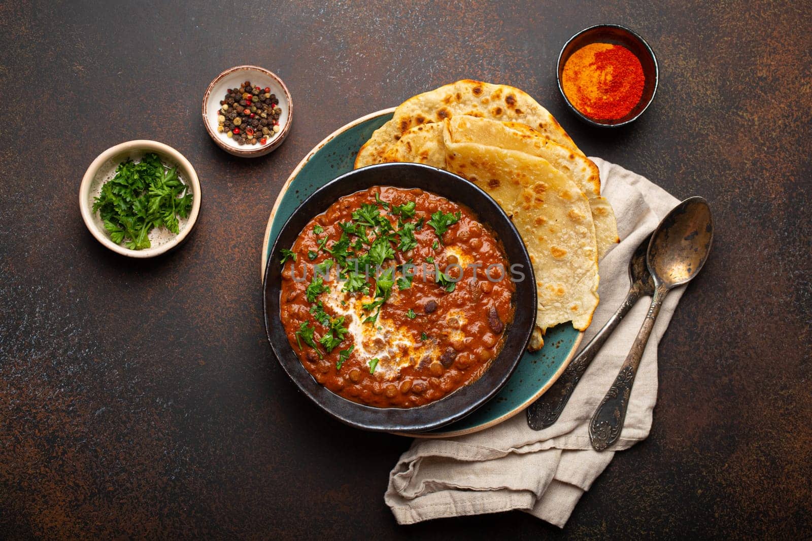 Traditional Indian Punjabi dish Dal makhani with lentils and beans in black bowl served with naan flat bread, fresh cilantro and two spoons on brown concrete rustic table top view by its_al_dente