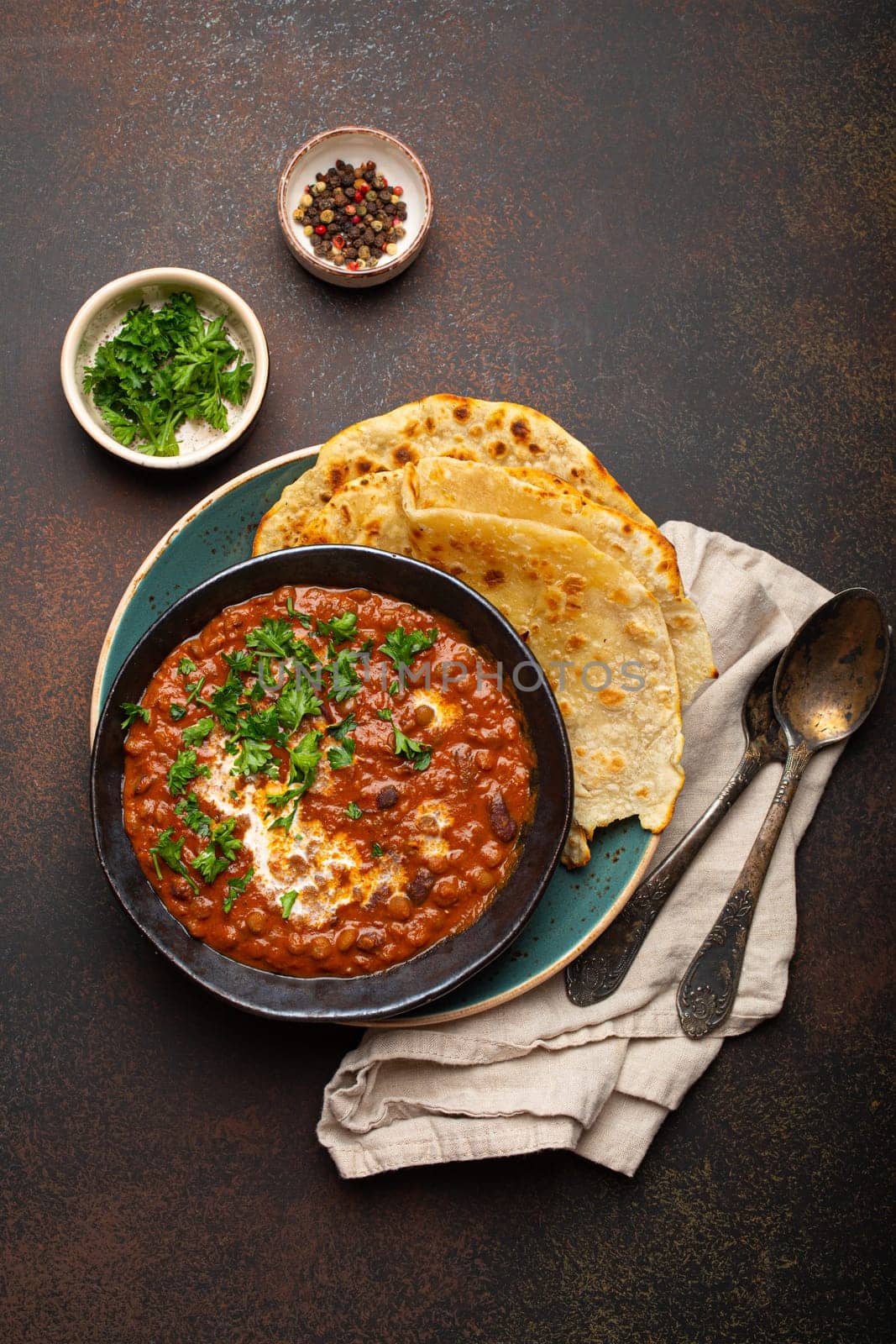 Traditional Indian Punjabi dish Dal makhani with lentils and beans in black bowl served with naan flat bread, fresh cilantro and two spoons on brown concrete rustic table top view by its_al_dente