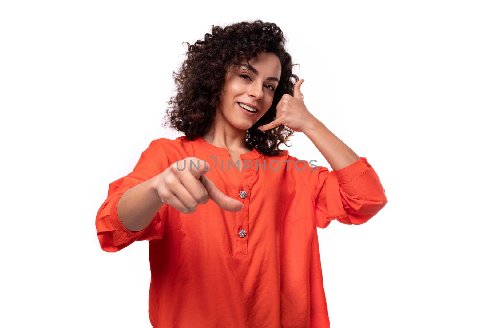 young caucasian lady with black curly hair dressed in an orange blouse points her finger at the camera.