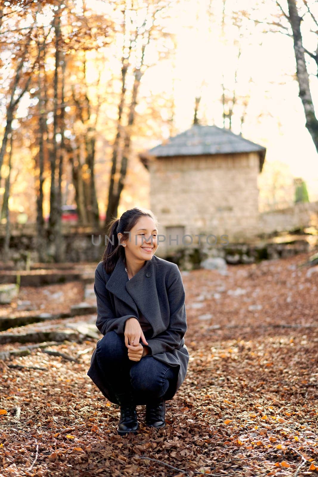 Young smiling woman squatting in autumn park and looking away. High quality photo