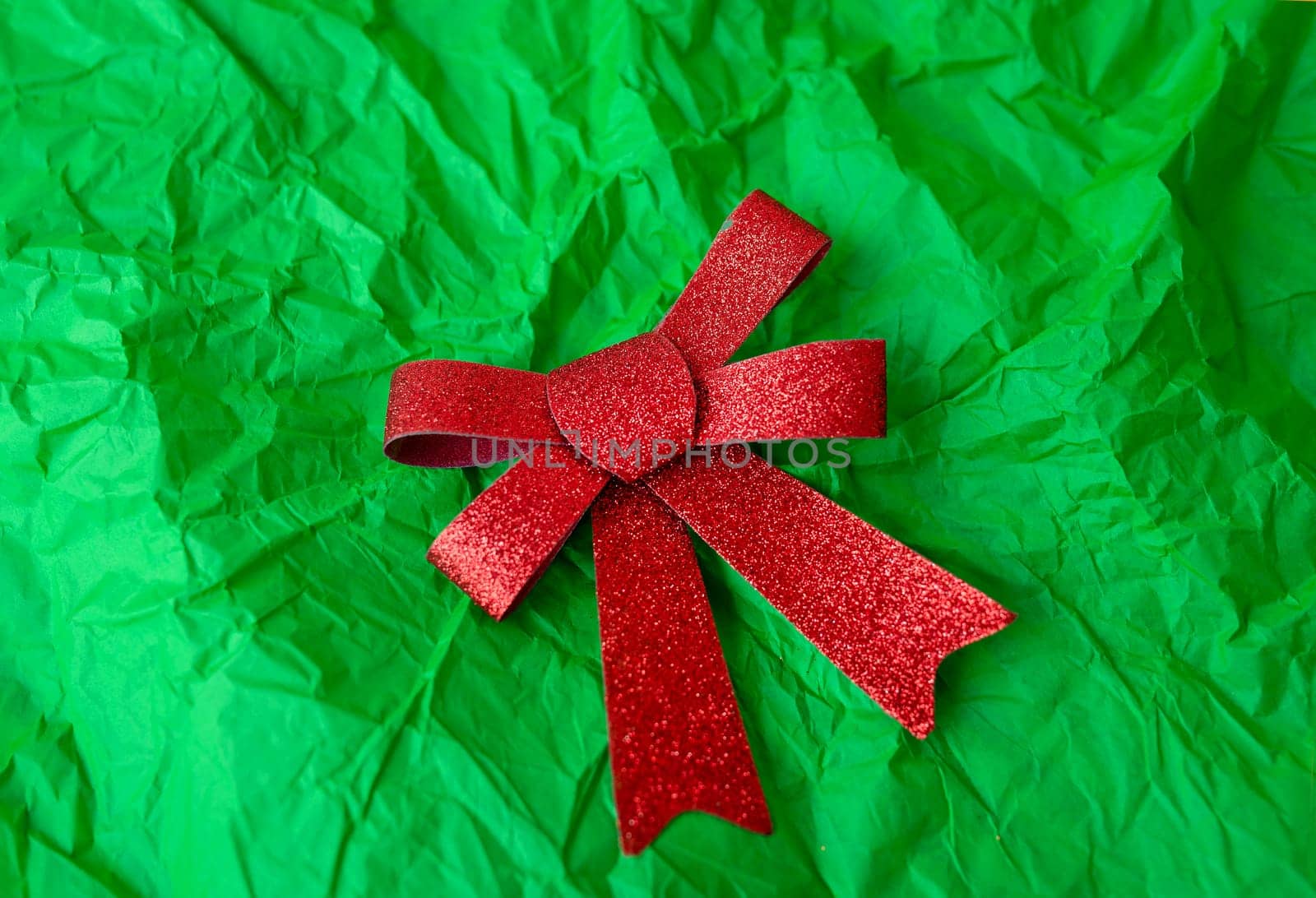 A photo of a red glittery bow on a green crumpled paper background. The bow is in the center of the image and the background fills the frame. by sfinks