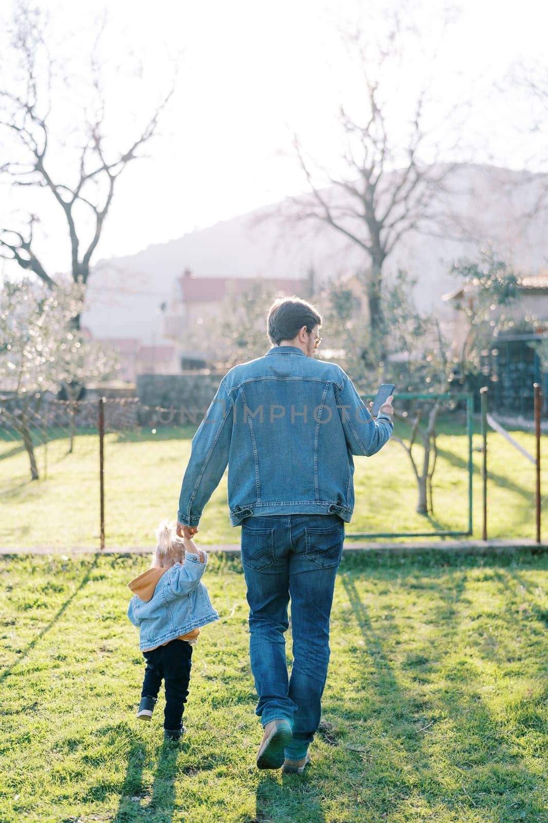 Dad leads a little girl by the hand through a green lawn to the house, looking at the phone. Back view by Nadtochiy