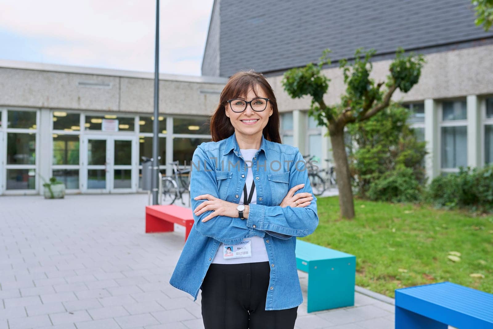 Middle aged confident woman teacher with crossed arms near school building by VH-studio