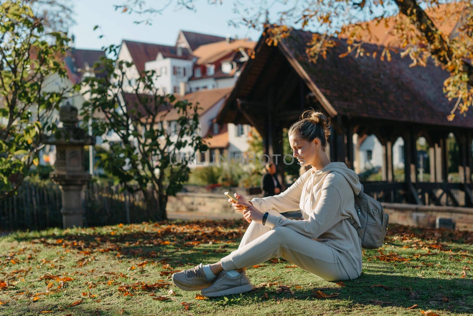 Young fashionable teenage girl with smartphone in park in autumn sitting at smiling. Trendy young woman in fall in park texting. Retouched, vibrant colors. Beautiful blonde teenage girl wearing casual modern autumn outfit sitting in park in autumn. Retouched, vibrant colors, brownish tones.