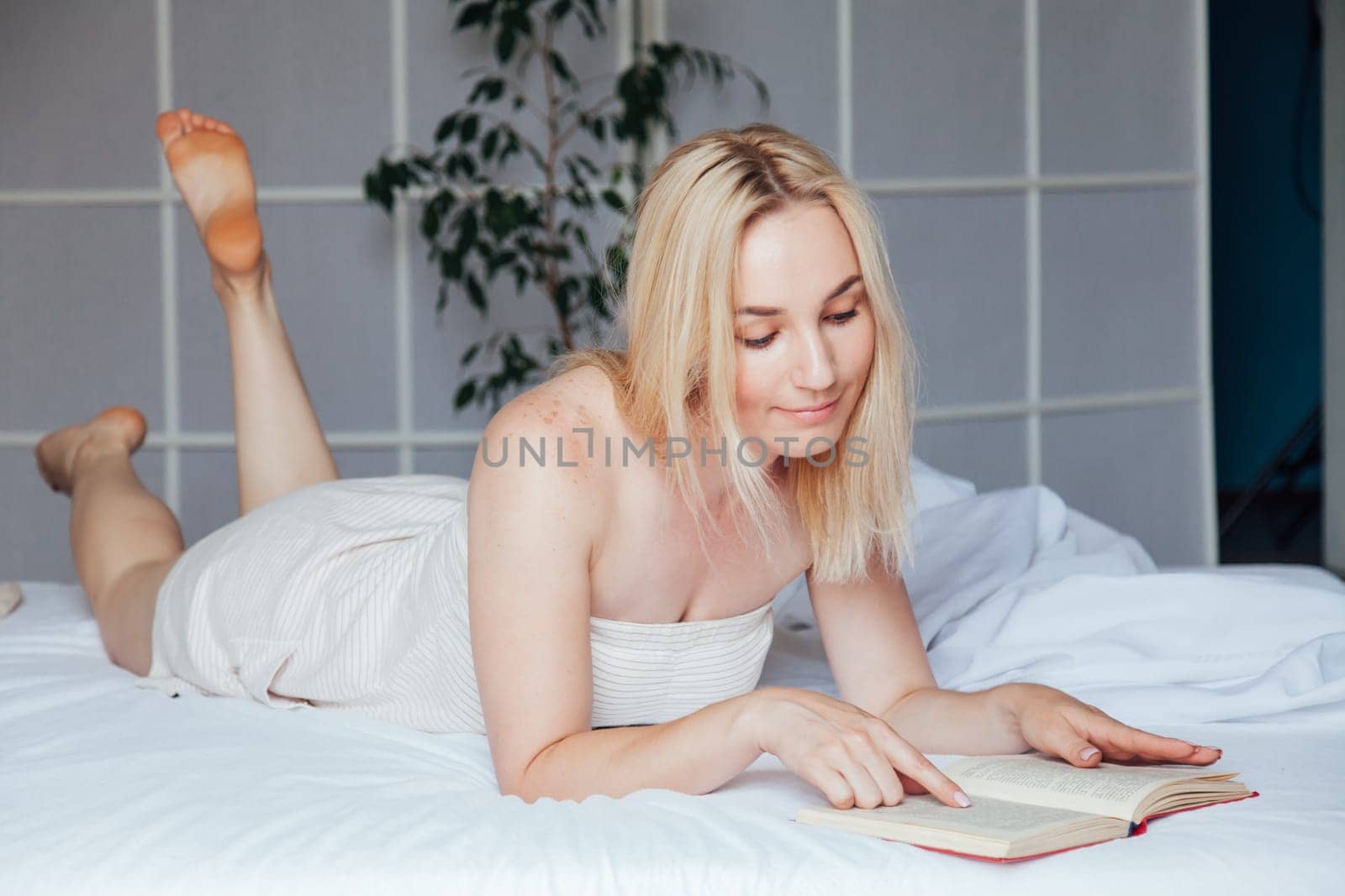 Beautiful blonde woman reads a book in the bedroom on the bed by Simakov