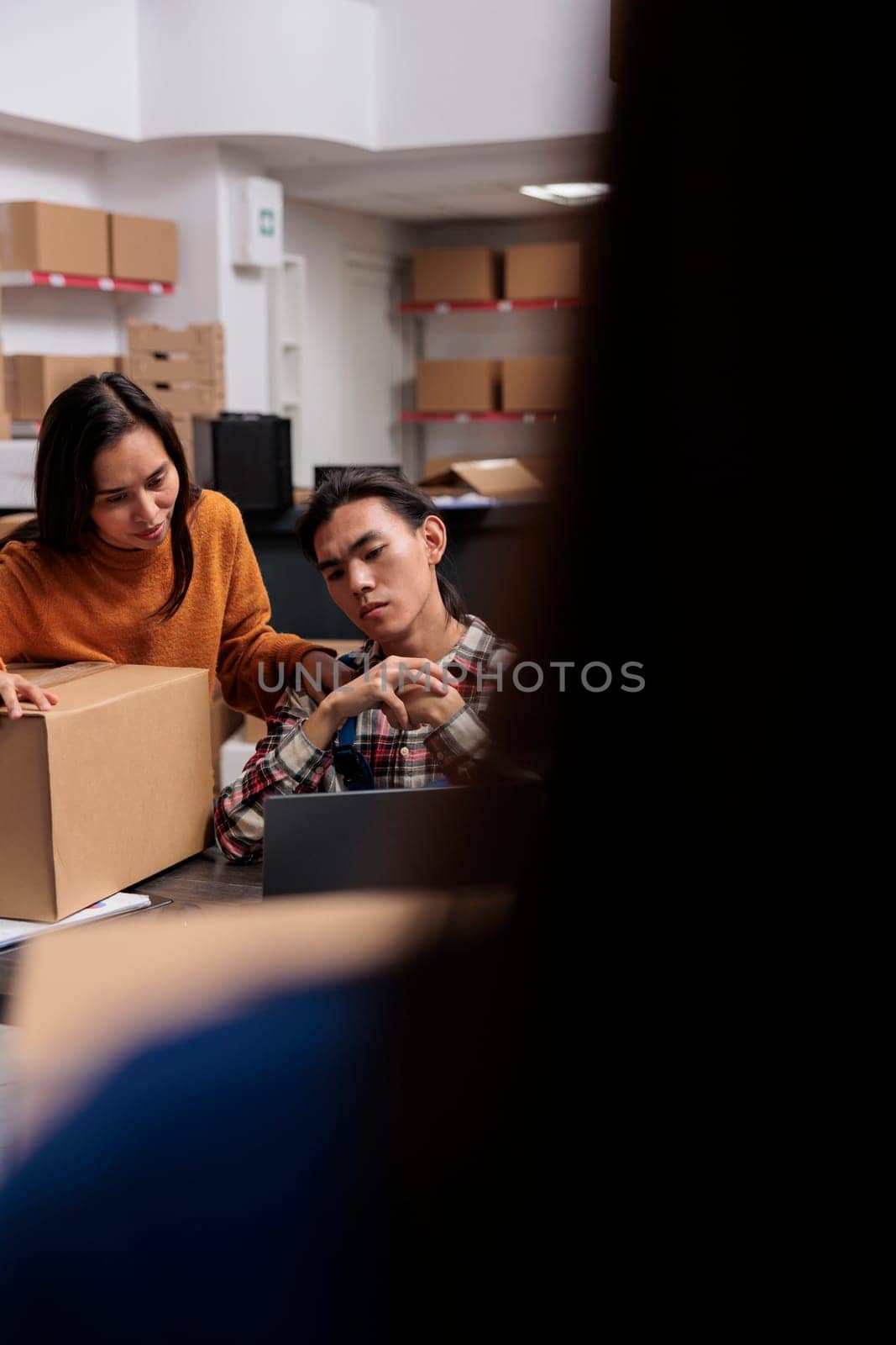 Warehouse order pickers asian team analyzing product information on laptop. Postal service storehouse employees managing parcel shipping route optimization on computer in storage room