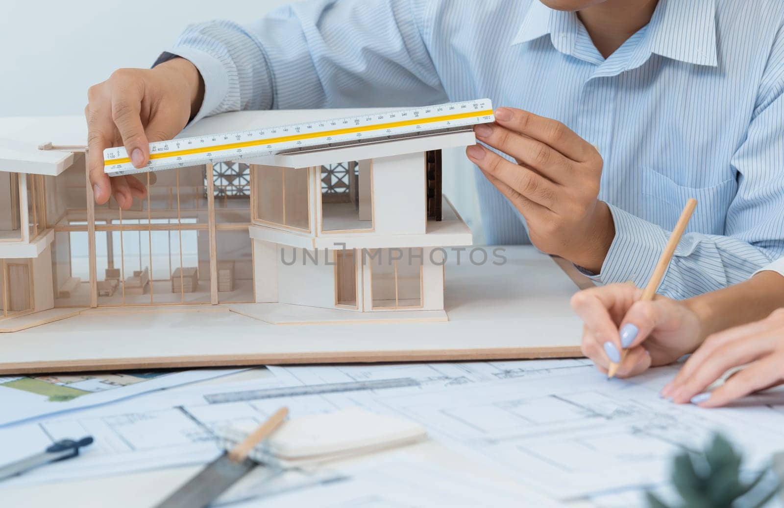 Professional male engineer hand measure house model by using ruler while beautiful cooperative coworker working together by writing on blueprint. Creative design and teamwork. Closeup. Immaculate.