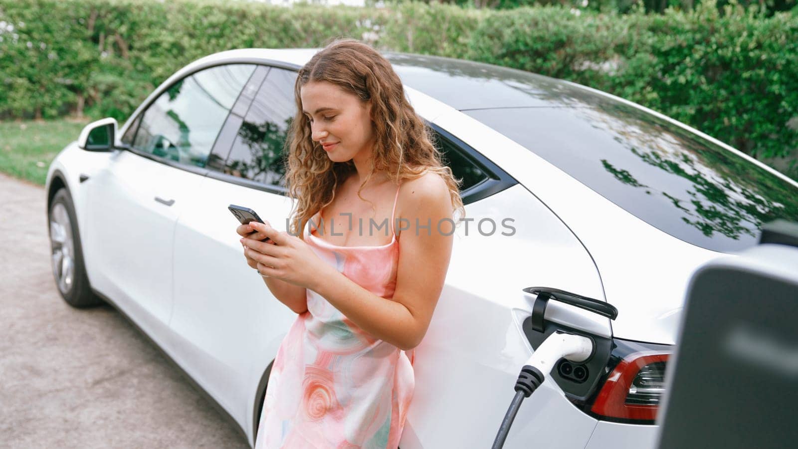 Young woman use smartphone to pay for electricity at public EV car charging station in nature. Modern environmental and sustainable automobile transportation lifestyle with EV vehicle. Synchronos