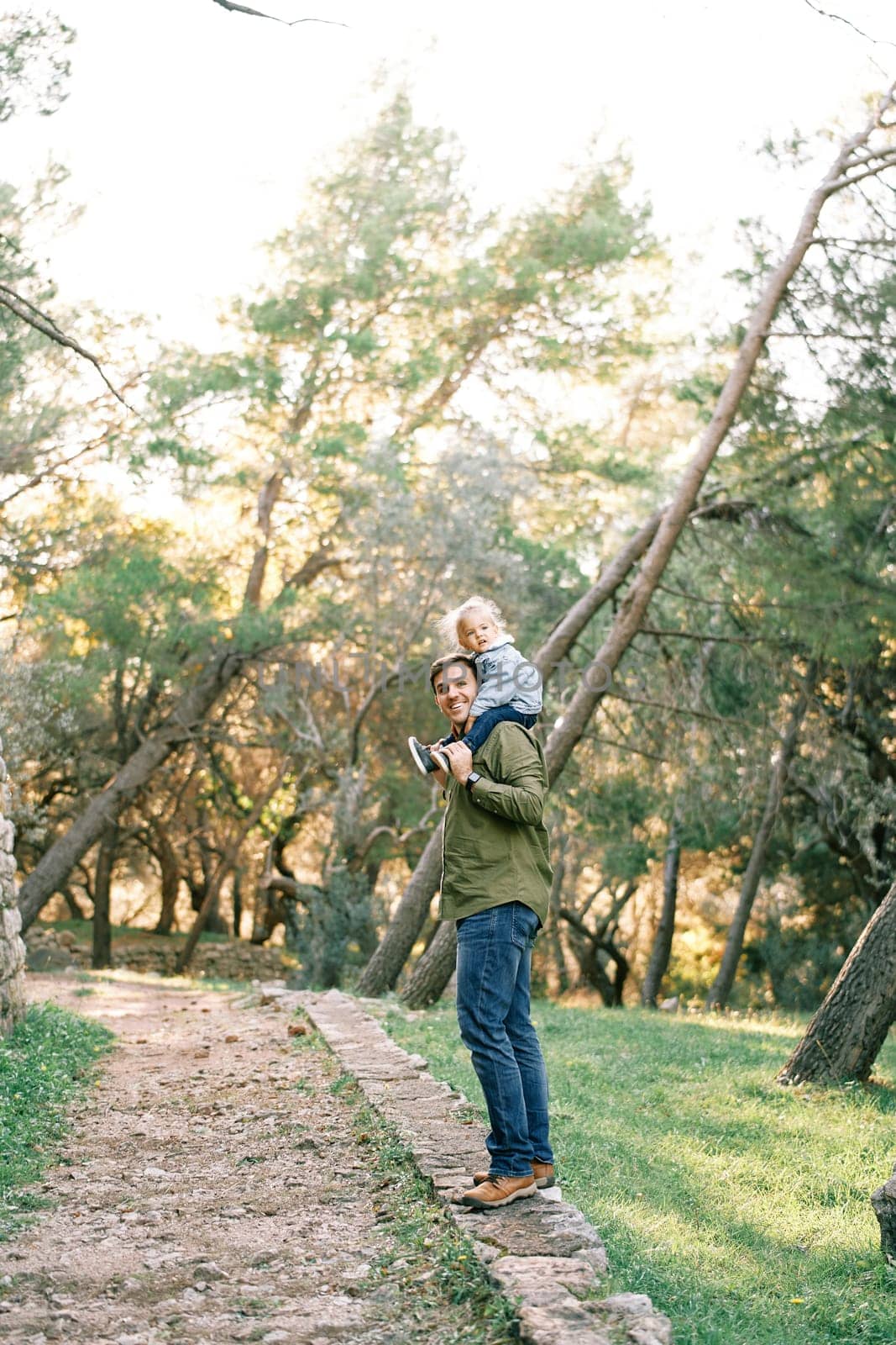 Smiling dad with a little girl on his shoulders stands half-turned on a paved path in the forest by Nadtochiy