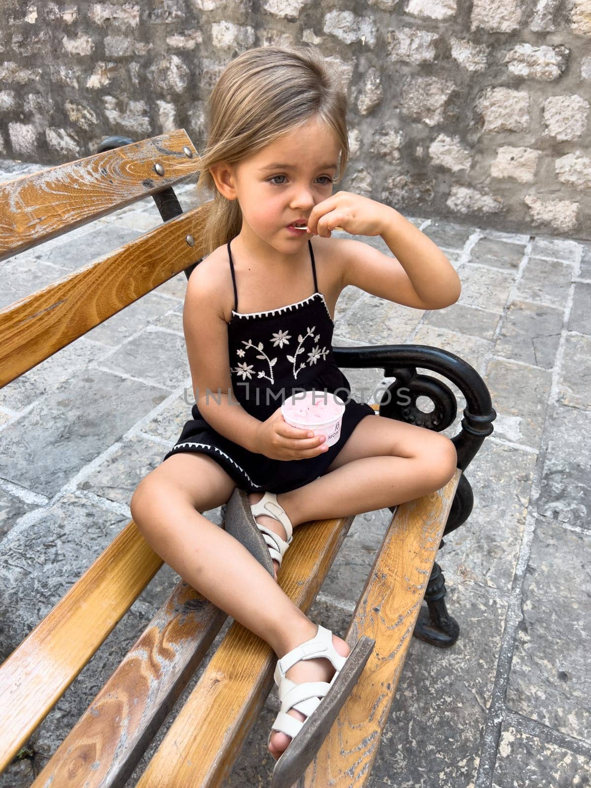 Little girl eats ice cream with a spatula from a cup while sitting on a bench by Nadtochiy