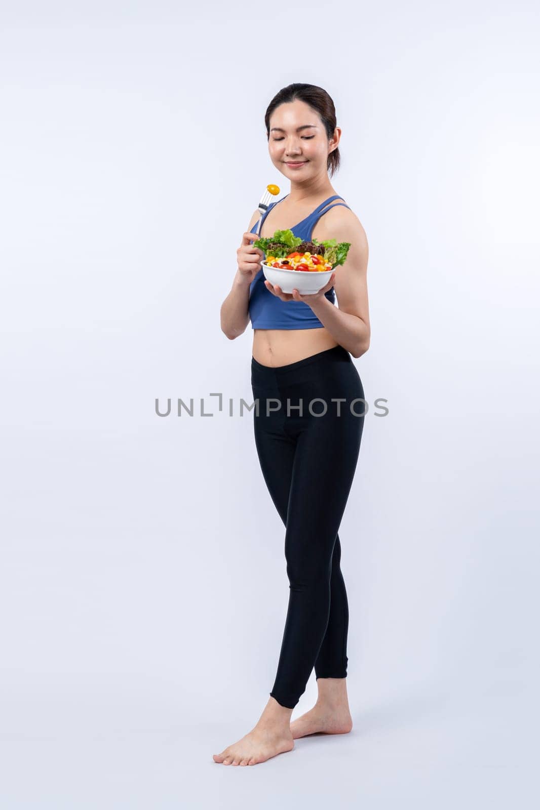 Young sporty Asian woman in sportswear holding salad bowl fill with vibrant of fruit and vegetable. Natural youthful and fit body lifestyle with balance nutrition on isolated background. Vigorous