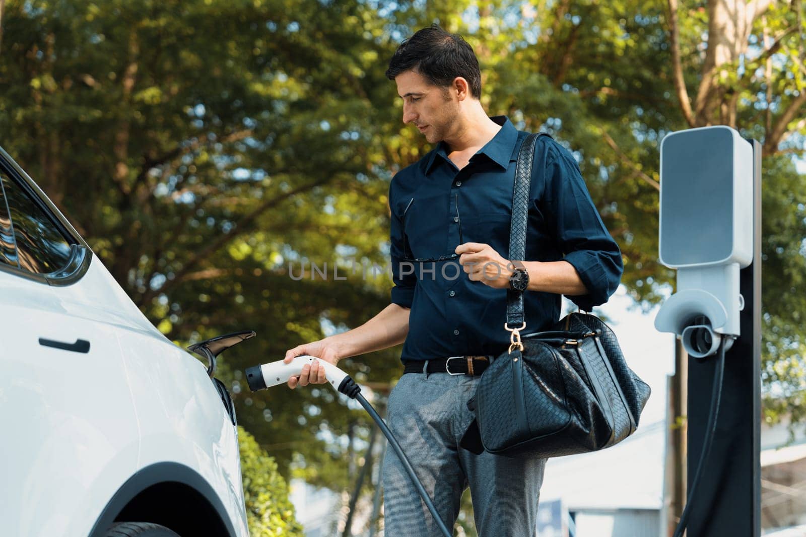 Young man recharge electric car's battery from charging station in outdoor green city park in springtime. Rechargeable EV car for sustainable environmental friendly urban travel lifestyle. Expedient