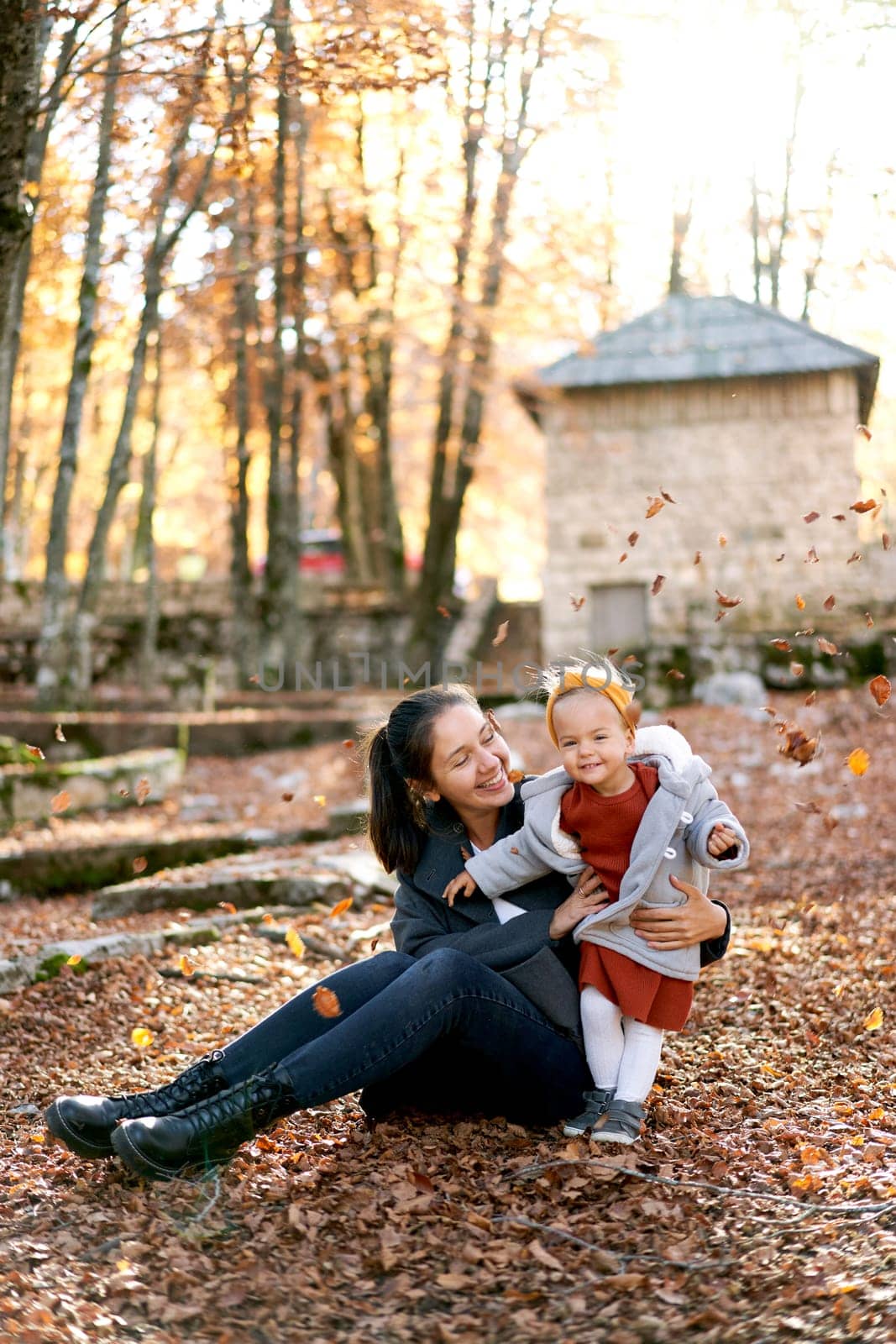 Smiling mother hugging little girl sitting on the ground in the park under falling leaves by Nadtochiy