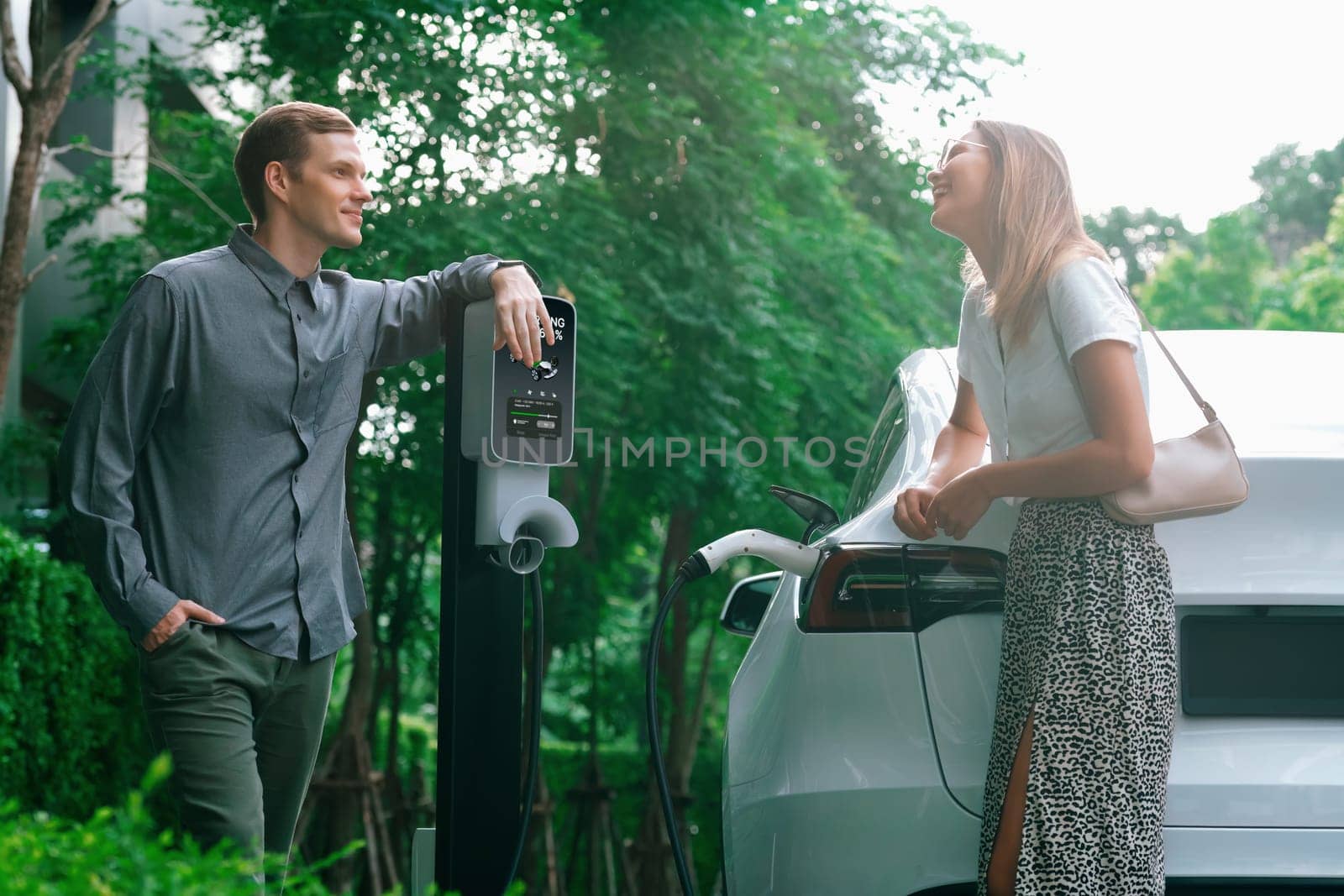 Young couple travel with EV electric car charging in green sustainable city outdoor garden in summer shows urban sustainability lifestyle by green clean rechargeable energy of electric vehicle innards