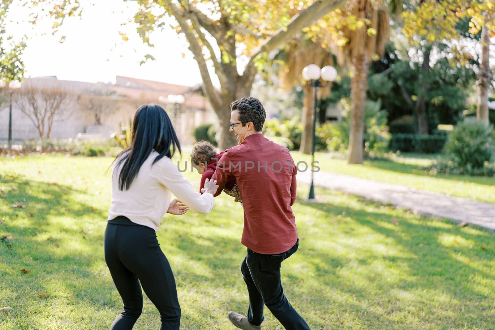 Laughing dad runs around mom in the park with a little girl in his arms. High quality photo