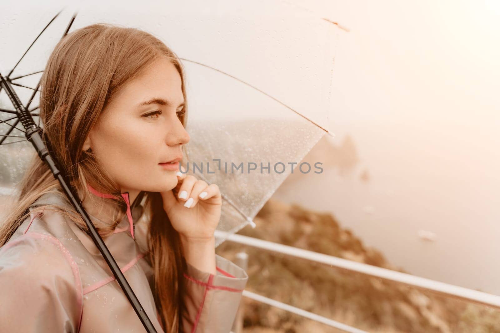 Woman rain park. Happy woman portrait wearing a raincoat with transparent umbrella outdoors on rainy day in park near sea. Girl on the nature on rainy overcast day