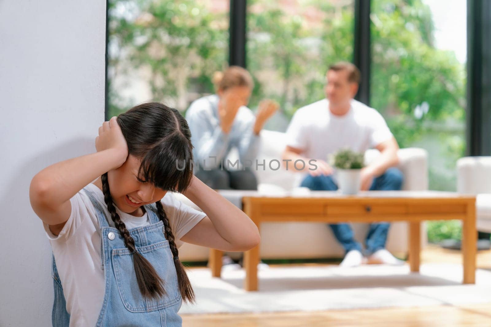 Stressed and unhappy girl huddle in corner, cover her ears with painful expression while her parent arguing in background. Domestic violence and traumatic childhood develop to depression. Synchronos