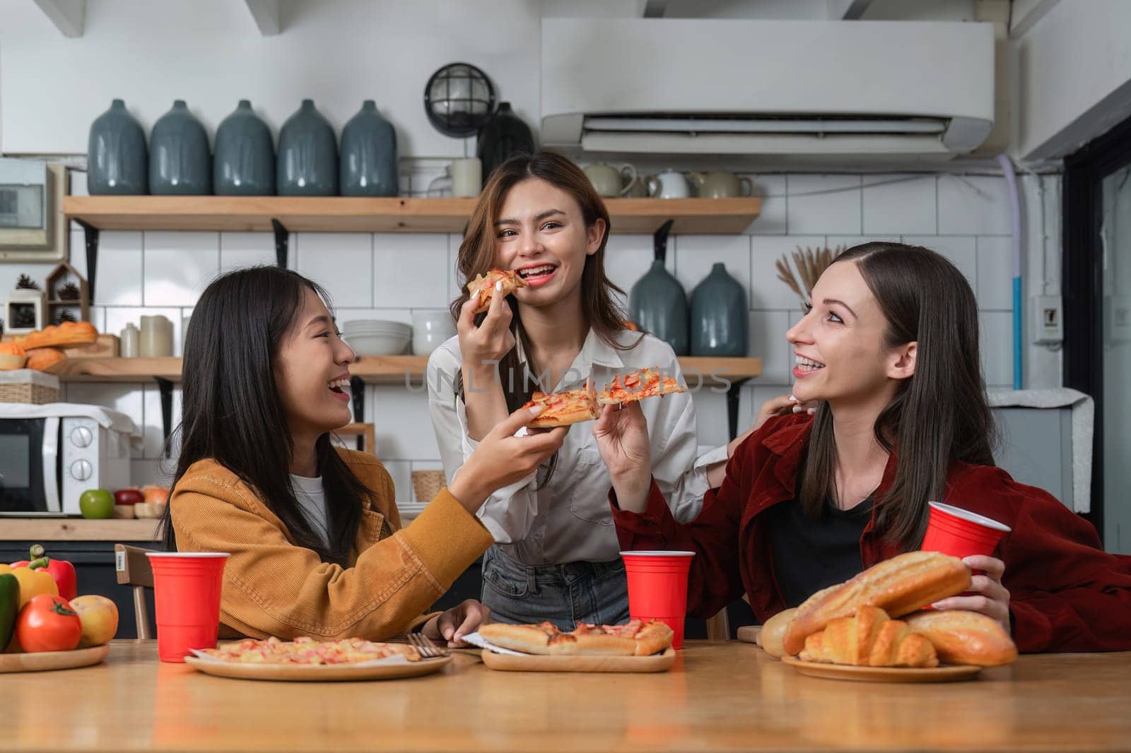 A group of young female friends have a party with pizza on the table and red drink glasses. Talk and live together happy, having fun at home..