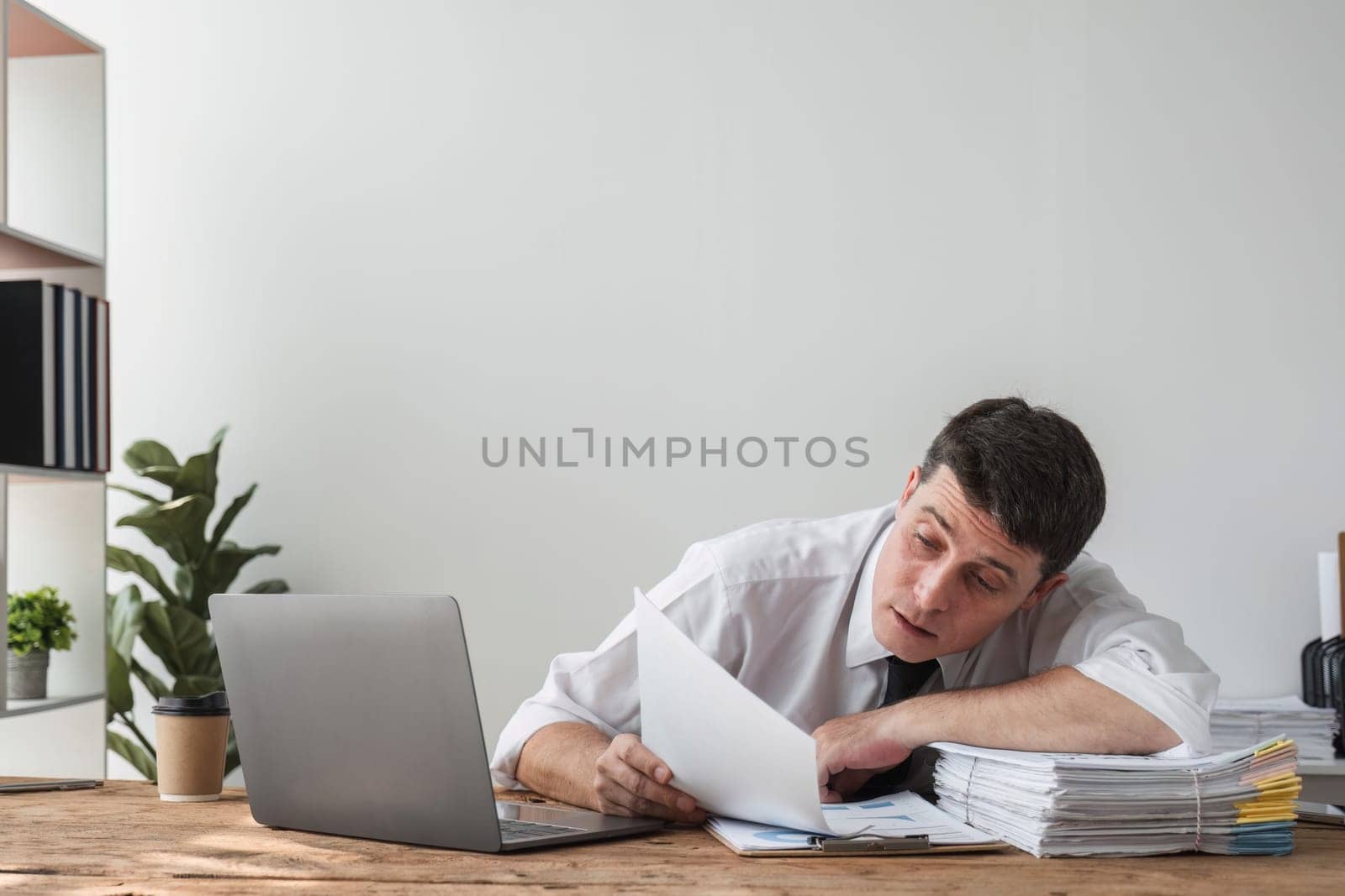 Unhappy young office worker feeling bored at work looking at laptop with a hopeless expression while sitting in the office. Male office worker feeling tired from monotonous work. by wichayada