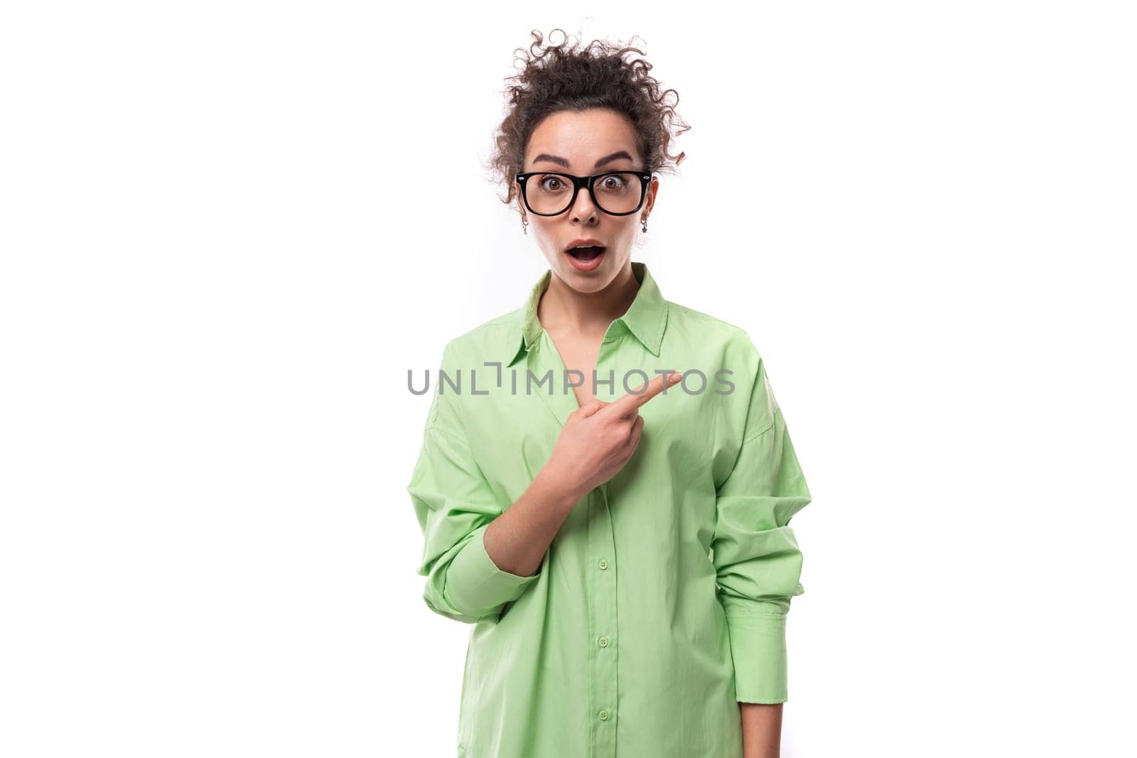 young surprised brunette businesswoman with curly hair dressed in a green shirt points her hand to the side.