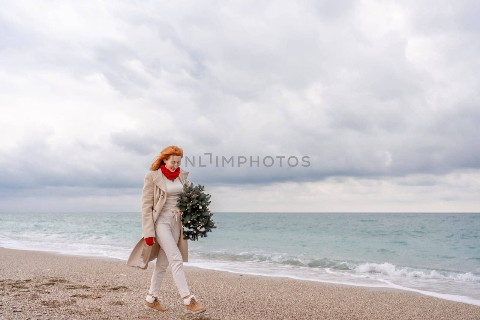 Redhead woman Christmas tree sea. Christmas portrait of a happy redhead woman walking along the beach and holding a Christmas tree in her hands. Dressed in a light coat, white suit and red mittens
