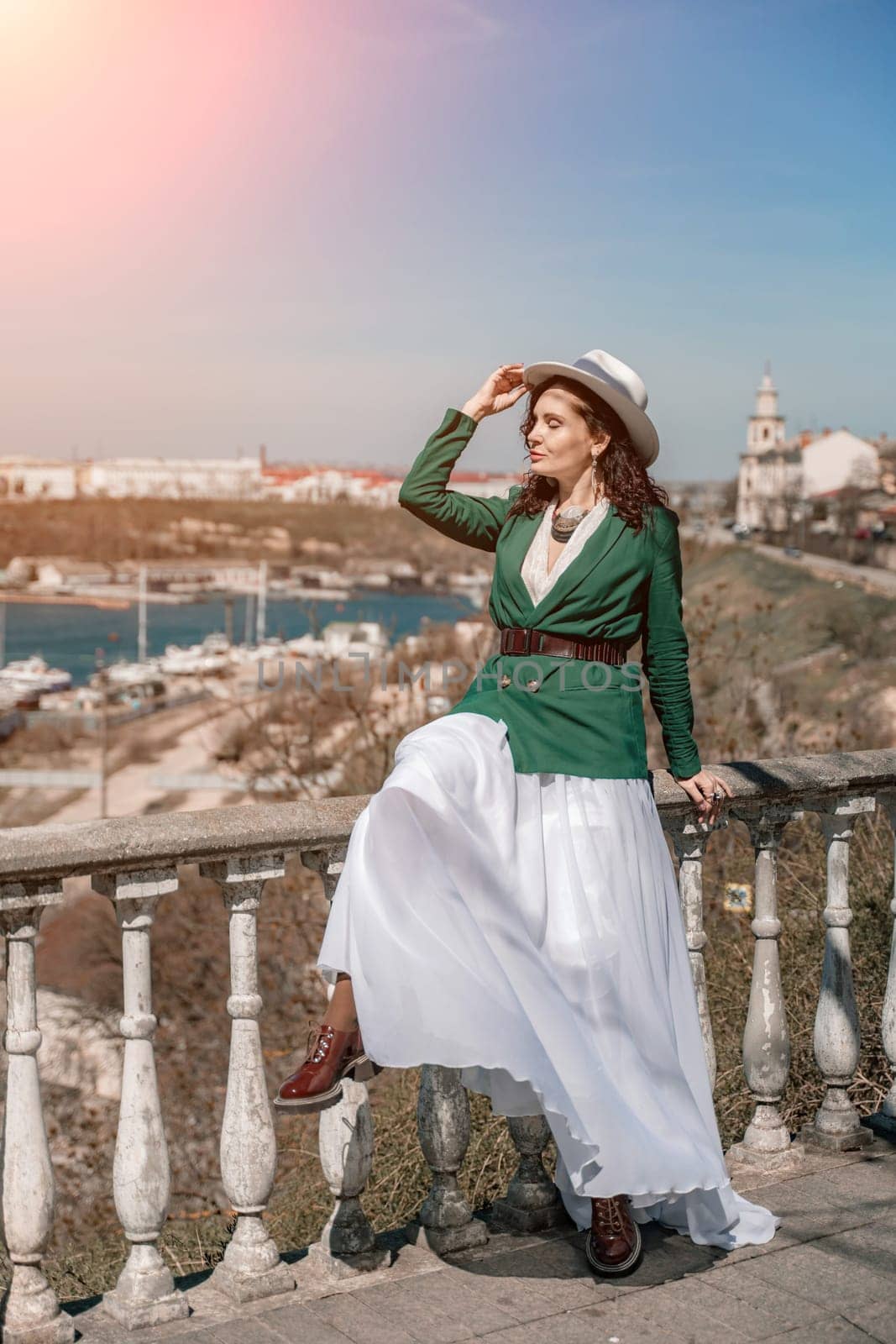 Woman walks around the city, lifestyle. A young beautiful woman in a green jacket, white skirt and hat is sitting on a white fence with balusters overlooking the sea bay and the city