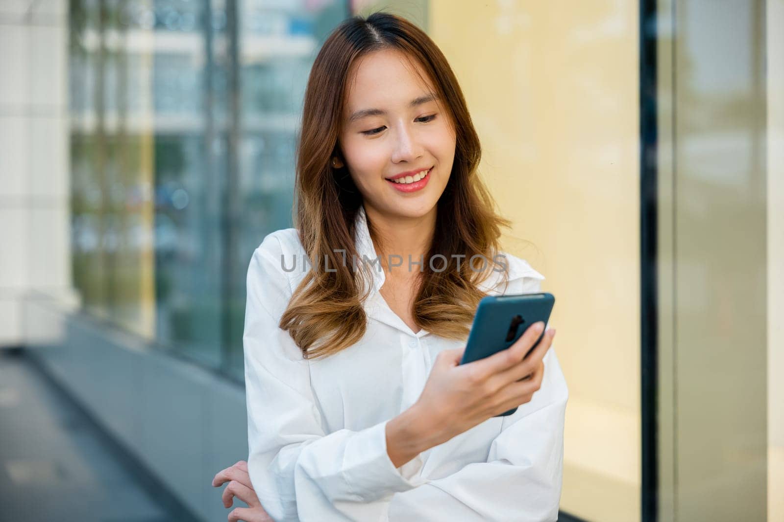 A smiling woman is using her smartphone outside by Sorapop